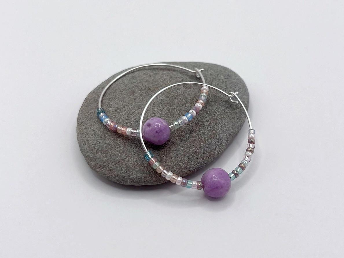 Close up of a pair of silver hoop earrings with lilac Jasper and seed beads laid on a purple pebble on a white background.