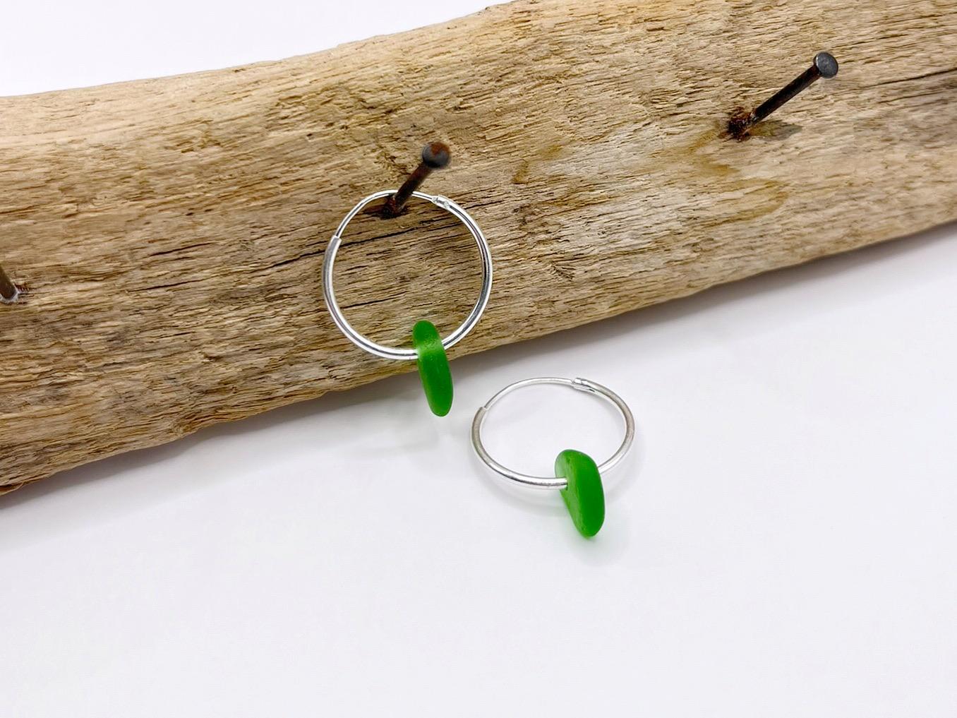 Silver hoop earrings with green sea glass hanging on a piece of driftwood.