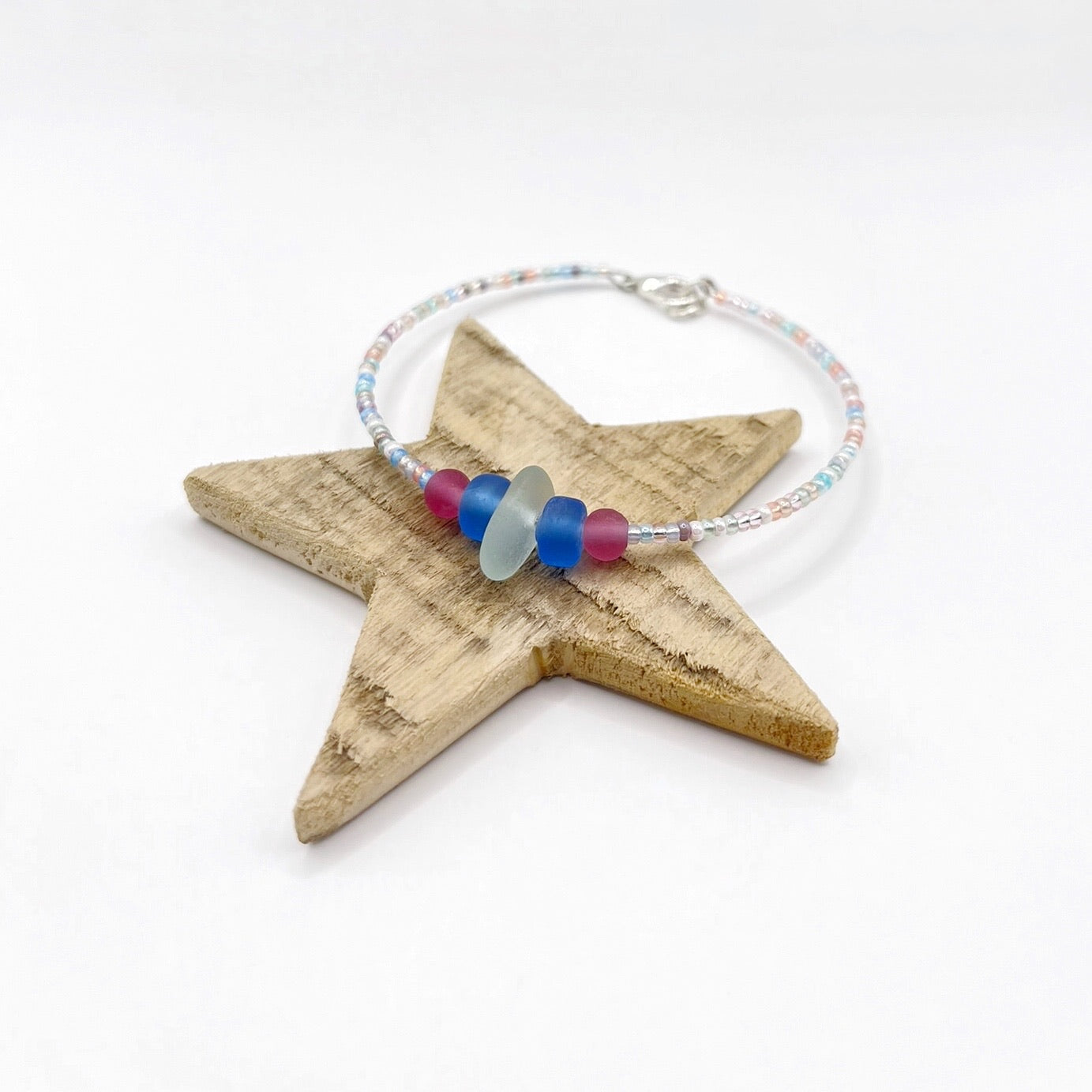 A pastel beaded bracelet with pink and blue frosted glass beads and a piece of sea foam coloured Cornish sea glass, finished with a silver plated lobster clasp and laid on a wooden star.