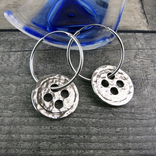 Silver hoop earrings with large round hammered silver buttons laid on a wooden surface.