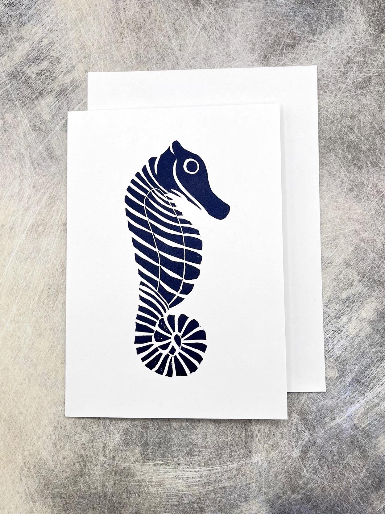 Original lino print of a navy blue seahorse design on a white card with matching envelope, laid on a textured background.