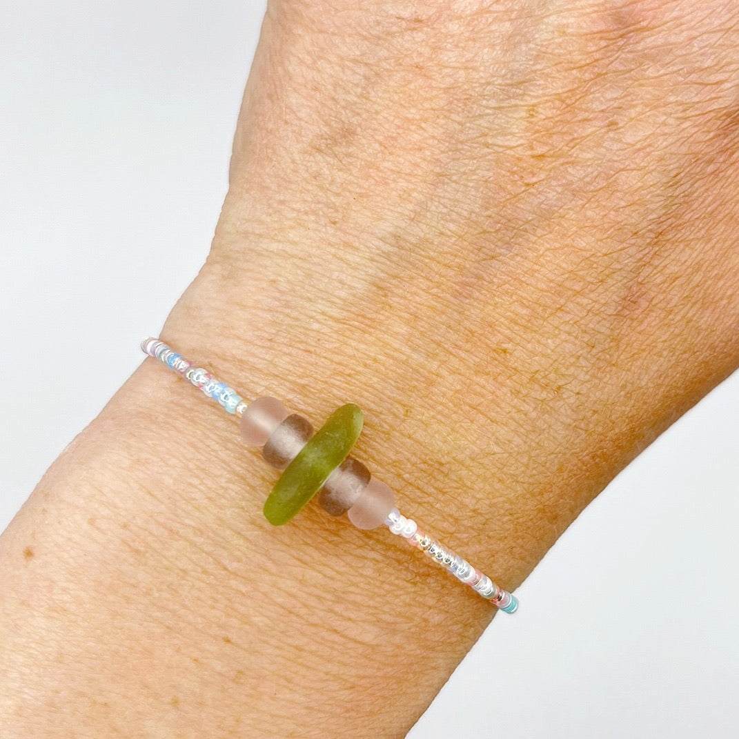 A pastel beaded bracelet with purple and lilac frosted glass beads and a piece of soft lime coloured Cornish sea glass, worn on a wrist.
