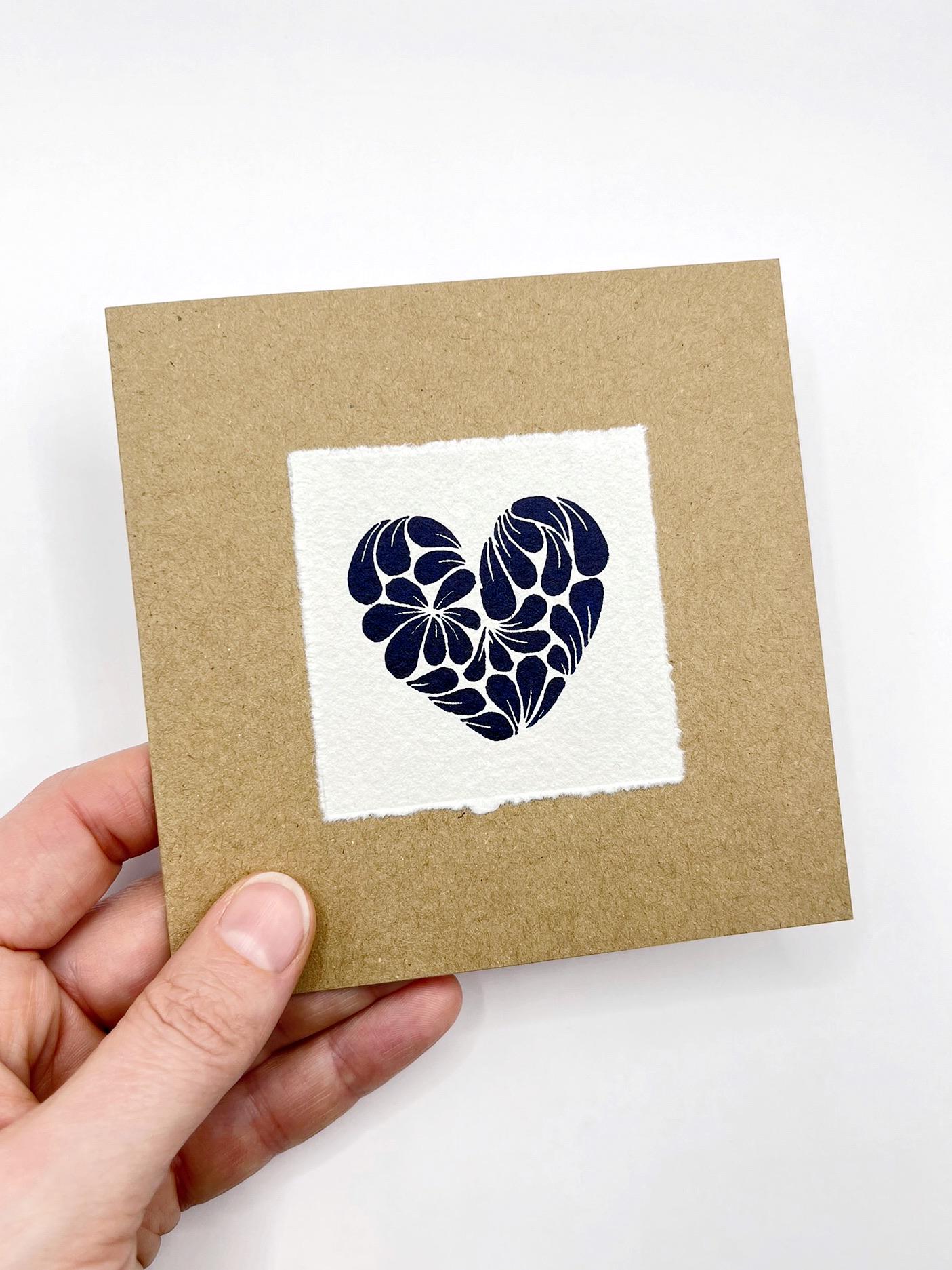 Hand holding a brown card with a white square featuring a blue floral heart design on a white background