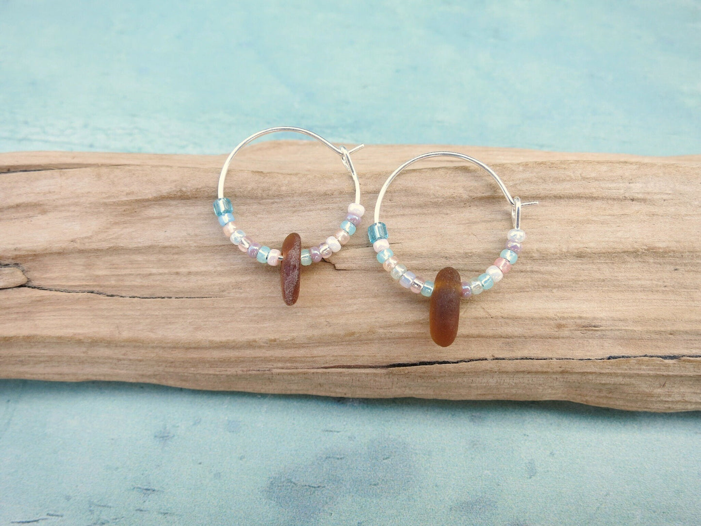 Silver hoop earrings with pastel seed beads and burnt orange sea glass laid on a piece of driftwood against a light blue background