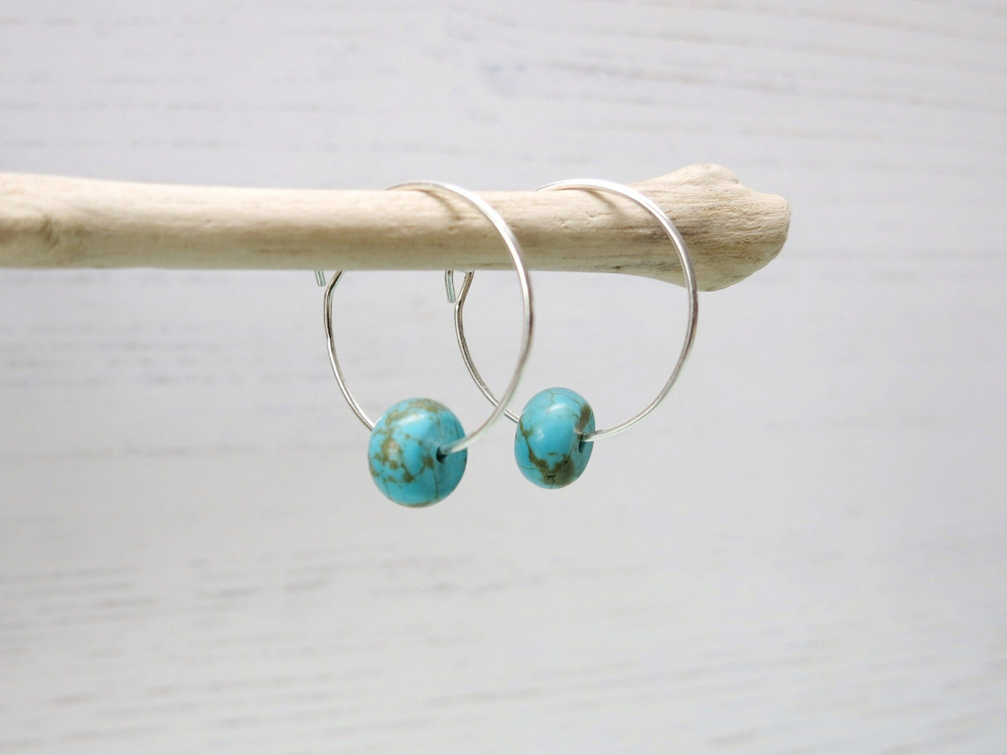 Silver hoop earrings with turquoise beads hanging on driftwood against a pale background