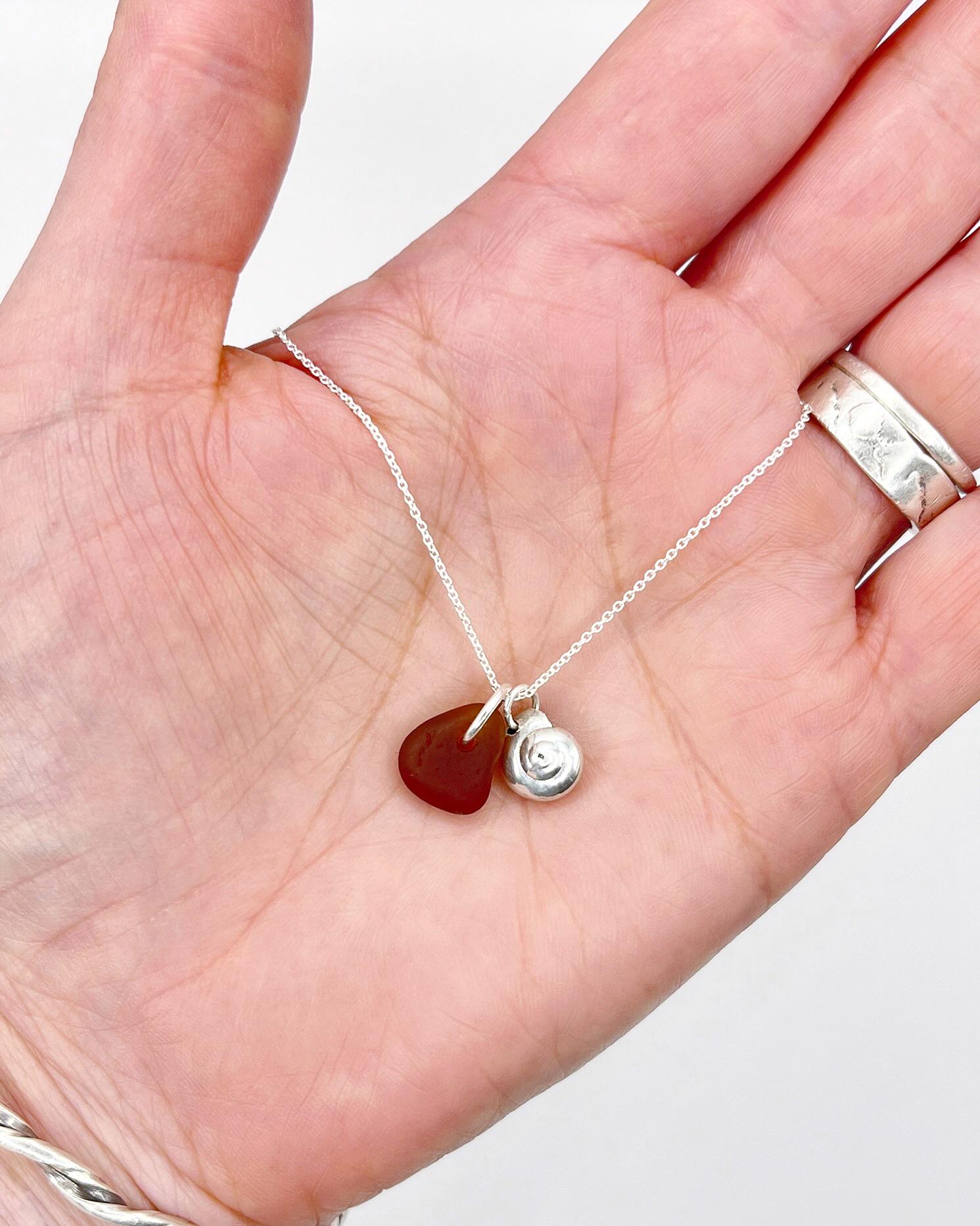 Hand holding a necklace with burnt orange sea glass and a silver shell pendant against a white surface