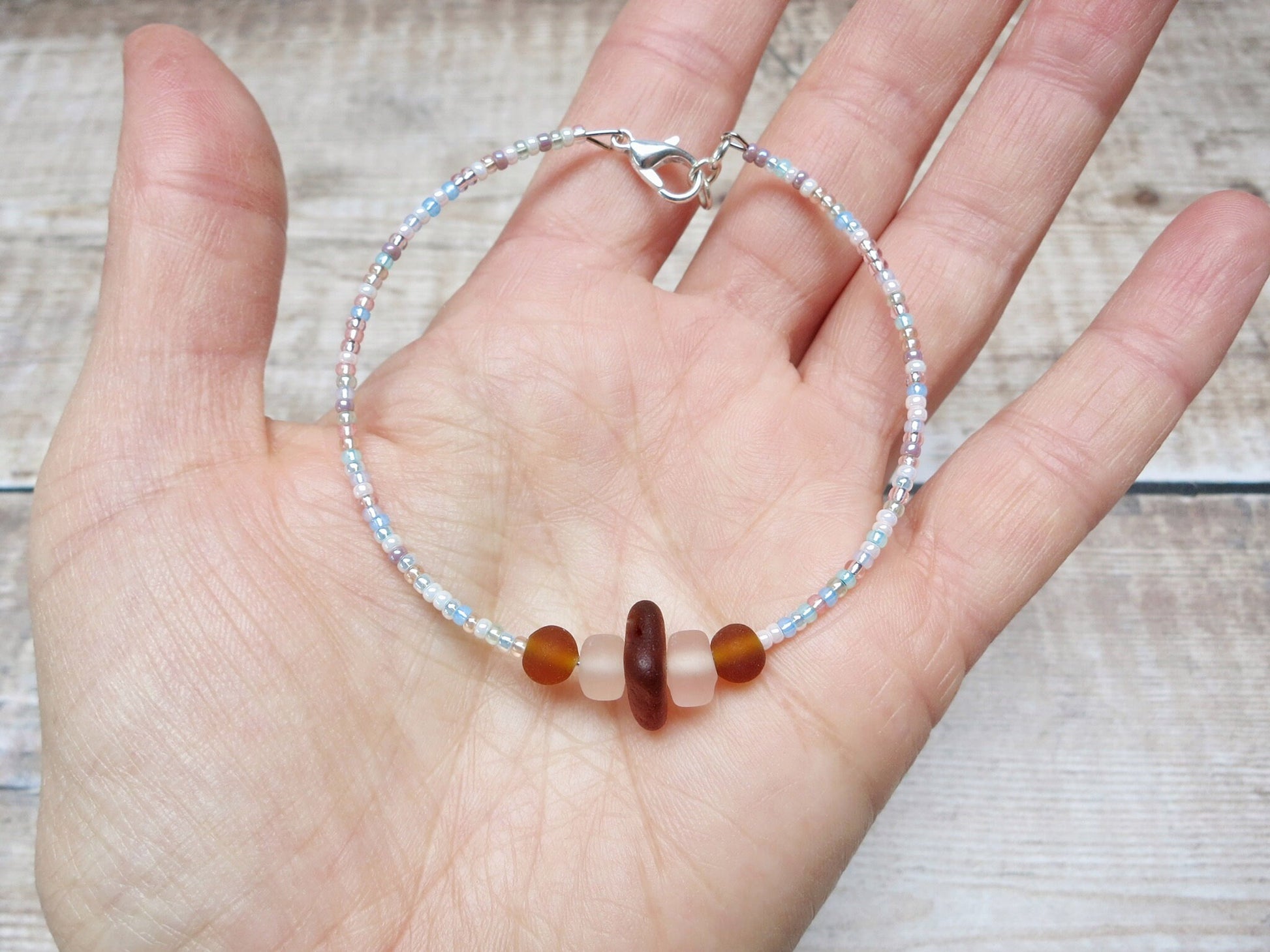 Hand holding bracelet with pastel seed beads and burnt orange sea glass against a textured background