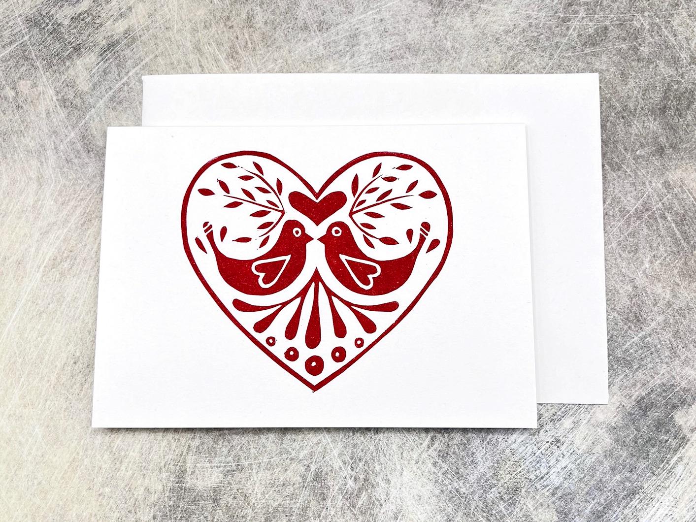 White card with an original lino printed red heart design featuring two birds laid on a grey textured background