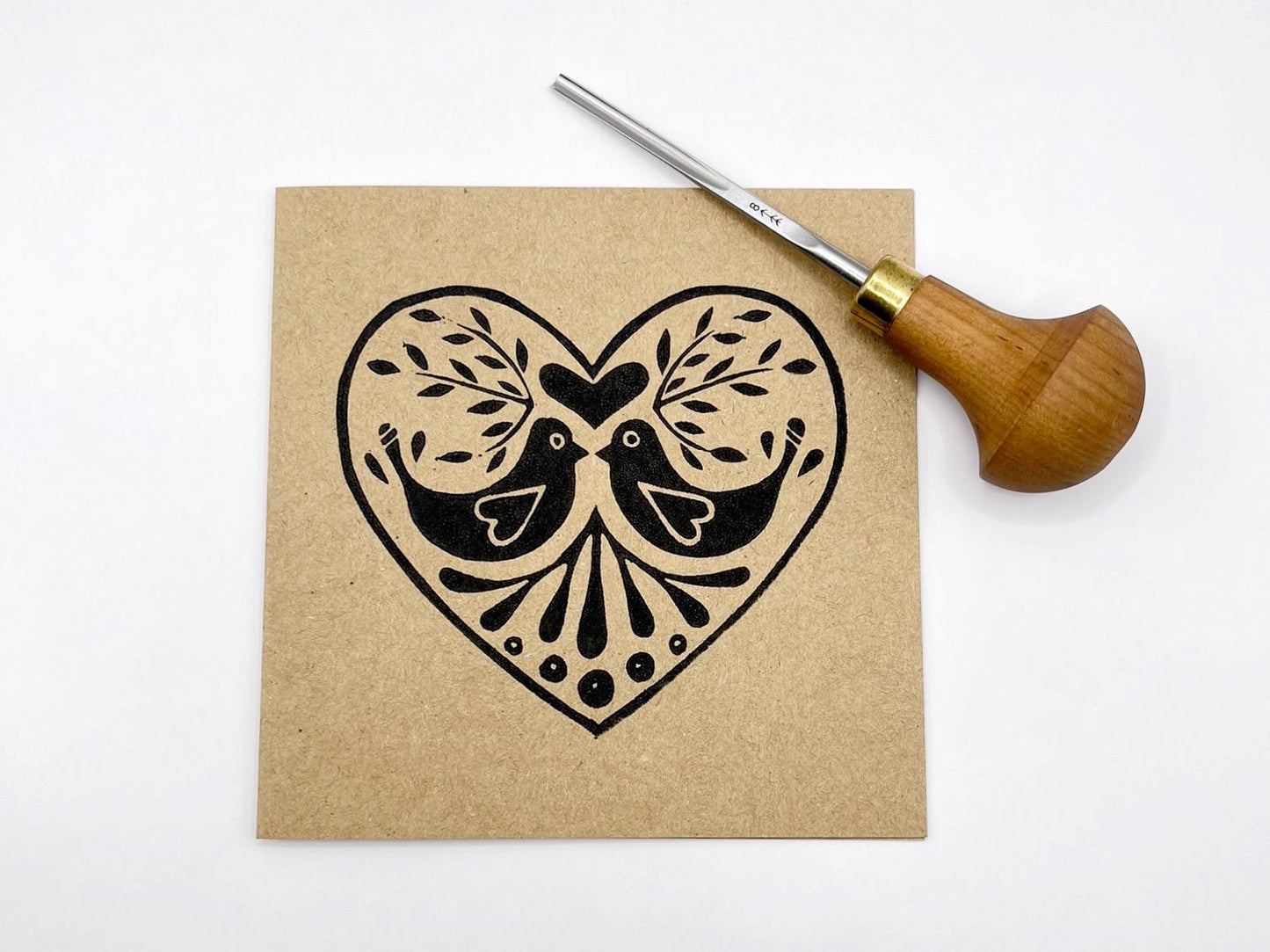 Kraft card with an original lino printed heart featuring two birds. Photo includes the lino cutting tool used against a white background.