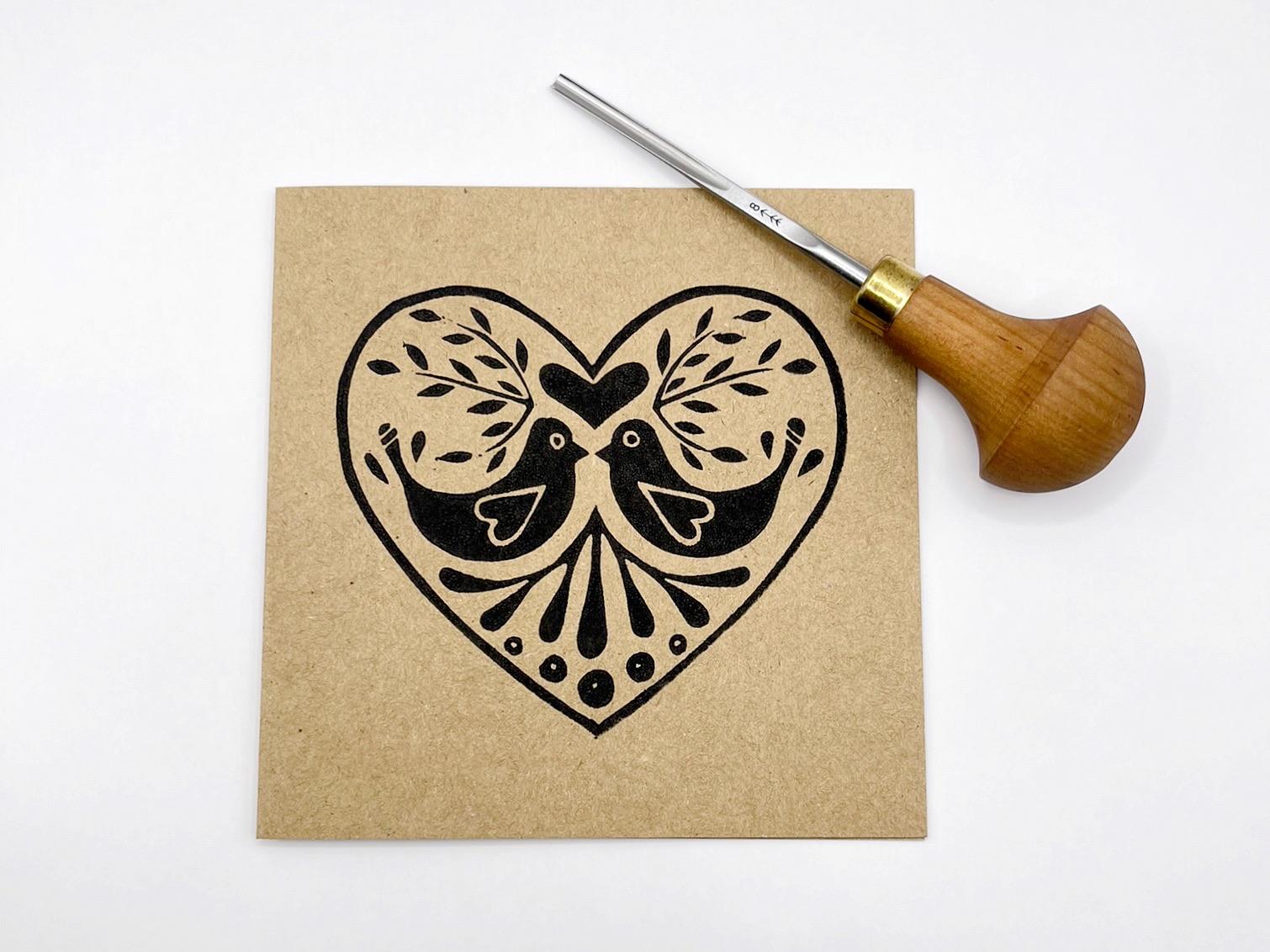 Kraft card with an original lino printed heart featuring two birds. Photo includes the lino cutting tool used against a white background.