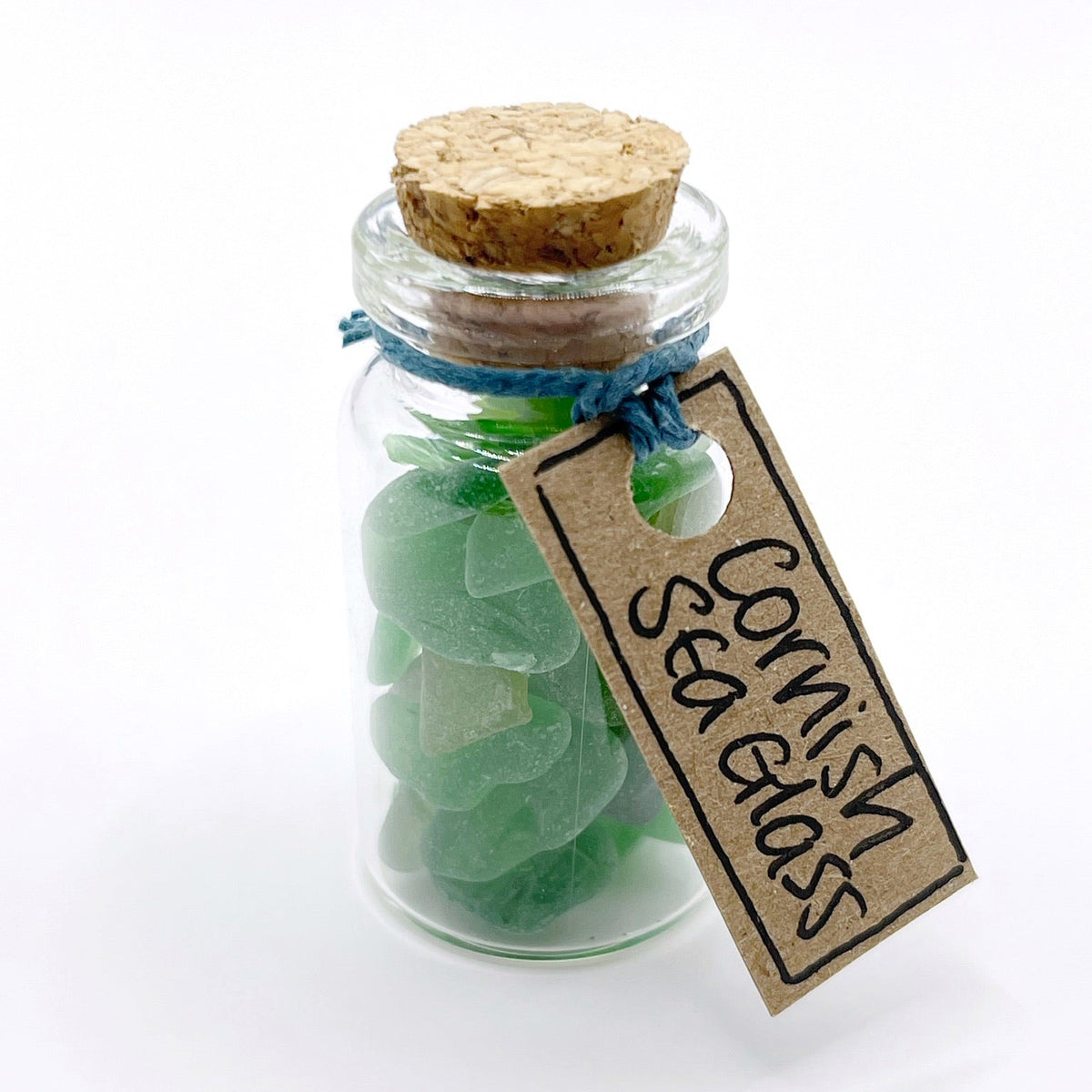 A small clear glass bottle with a cork stopper, filled with green sea glass pieces and tied with blue string. A kraft paper tag hangs from the neck and reads 'Cornish Sea Glass'.