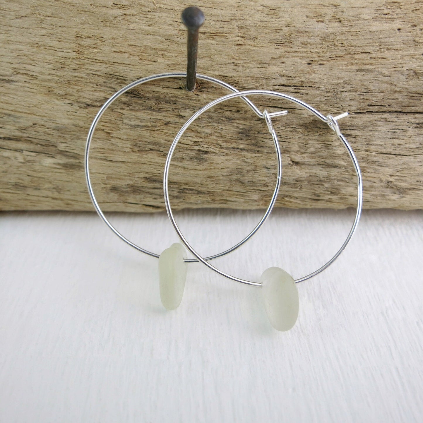 Silver hoop earrings with white sea glass hanging on driftwood against a white surface