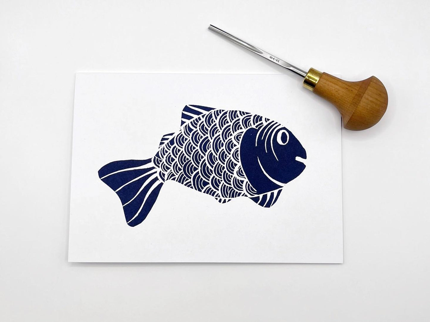 White card with an original lino printed blue fish, laid on a white background. Photo includes the lino cutting tool used.