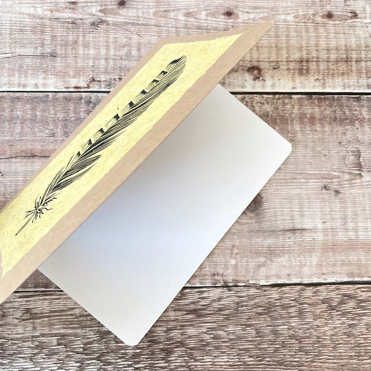 A5 kraft paper notebook or journal with a textured yellow handmade paper panel featuring a black lino ink feather illustration, photographed shown partially open on a rustic wooden plank background.