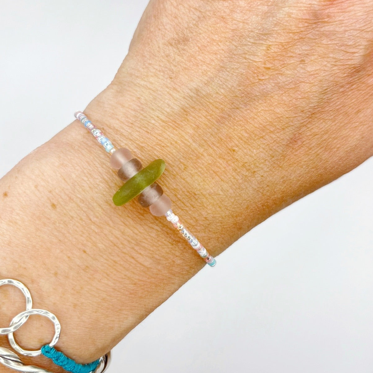 A pastel beaded bracelet with purple and lilac frosted glass beads and a piece of soft lime coloured Cornish sea glass, worn on a wrist.