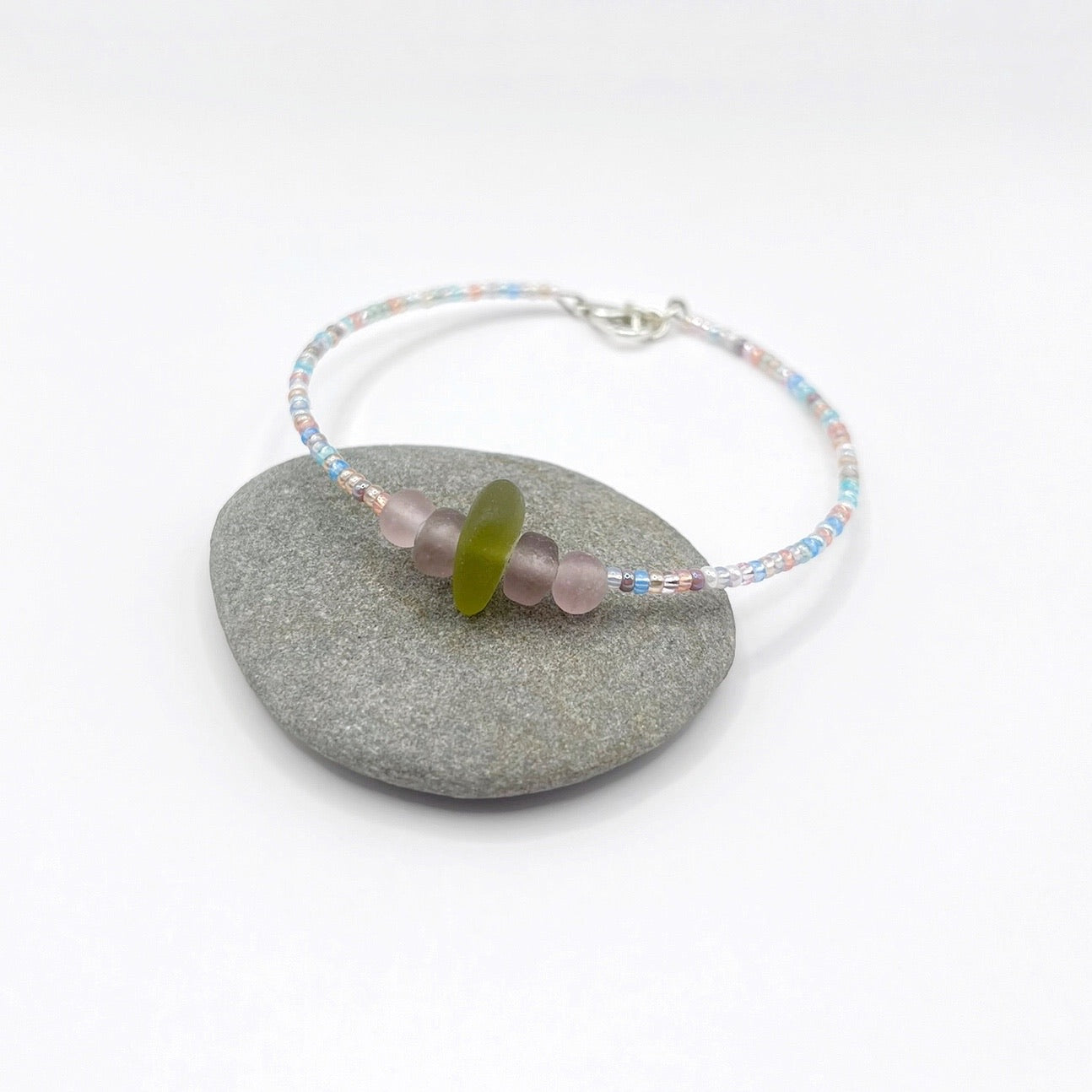 A pastel beaded bracelet with purple and lilac frosted glass beads and a piece of soft lime coloured Cornish sea glass, finished with a silver plated lobster clasp and laid on a grey pebble.
