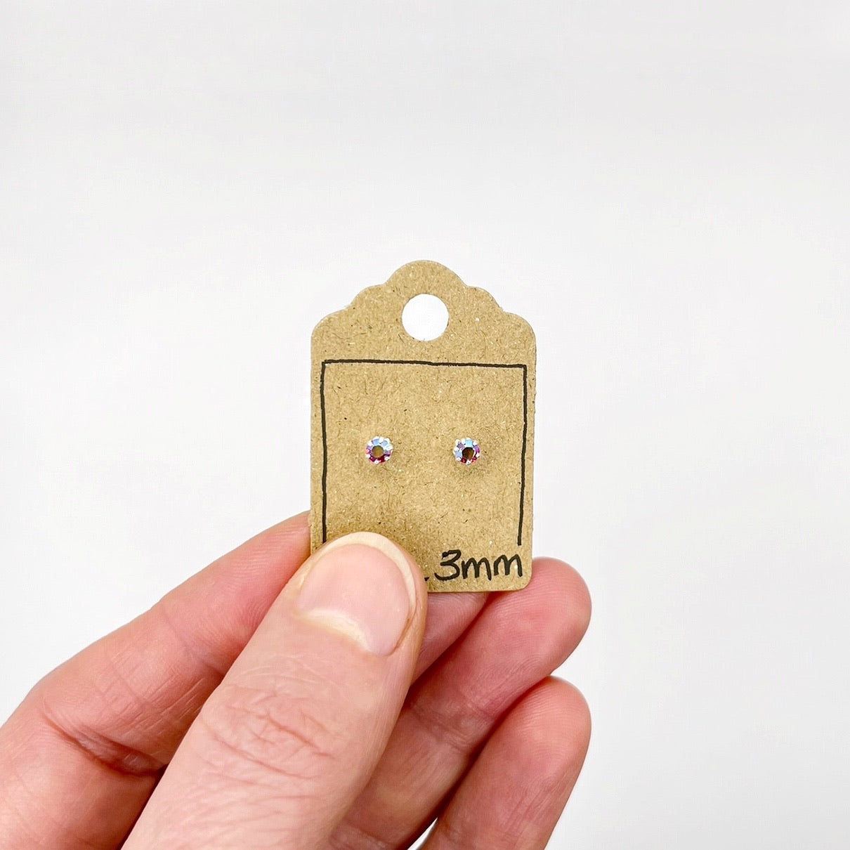 Hand holding a small cardboard earring display card with sparkly studs and '3mm' text on a white background