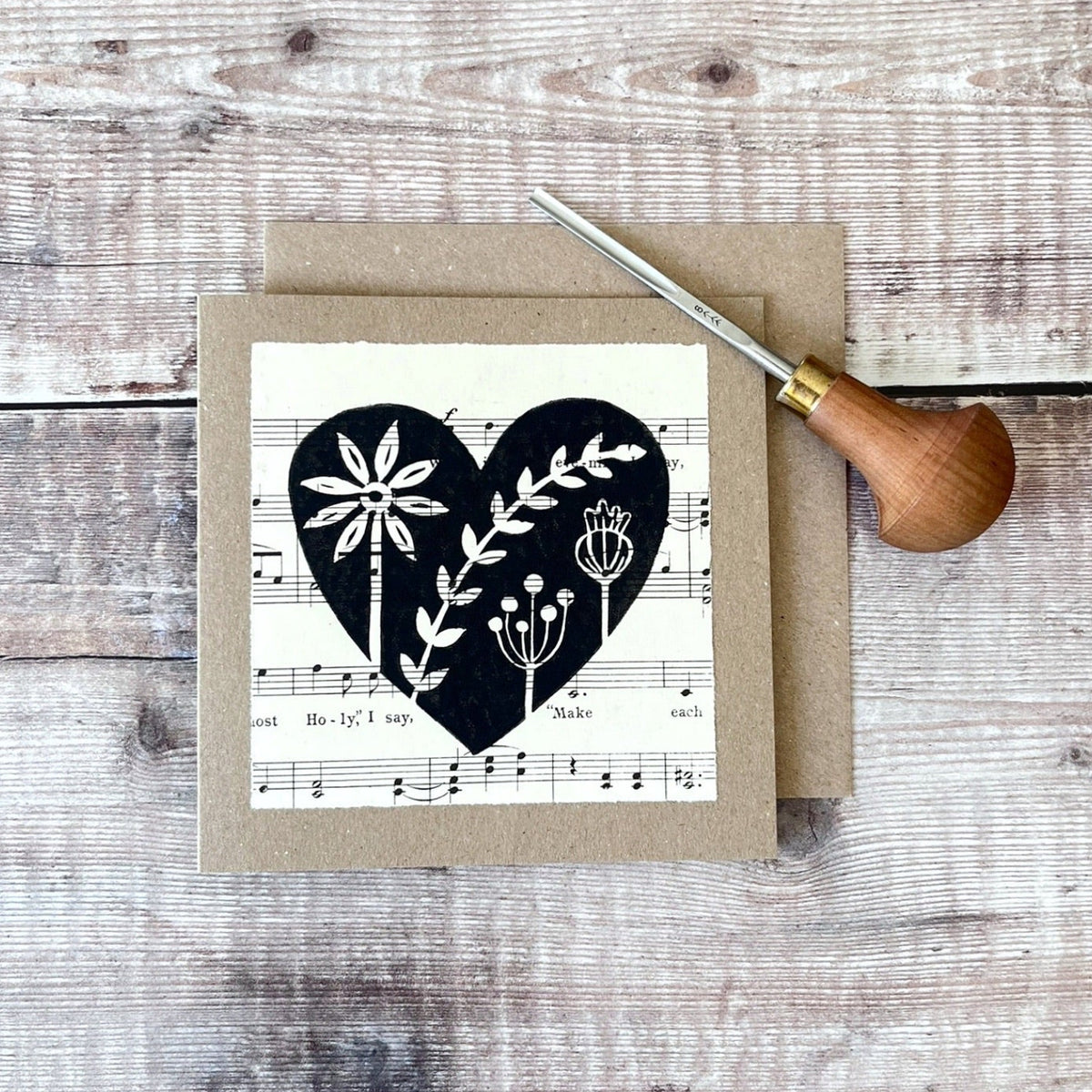 Square kraft greeting card with a black floral heart lino print on vintage music paper mounted on the front. Laid on a matching envelope on a wooden background. Photo includes the lino cutting tool used.
