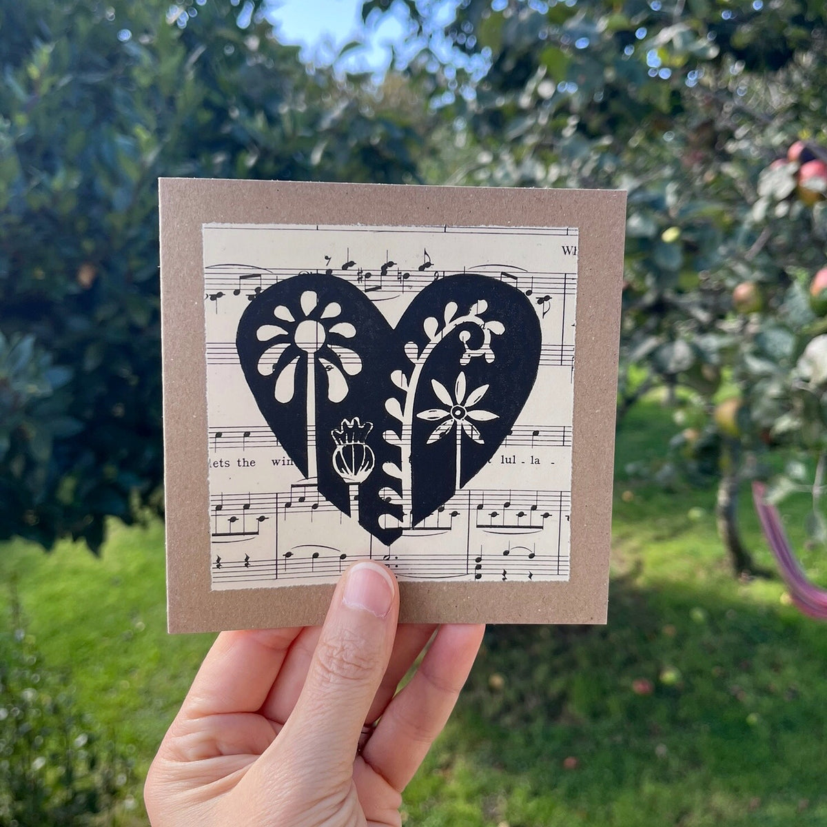 A hand holding a square kraft greeting card with a black floral heart lino print on vintage music paper mounted on the front. Held against a garden backdrop.