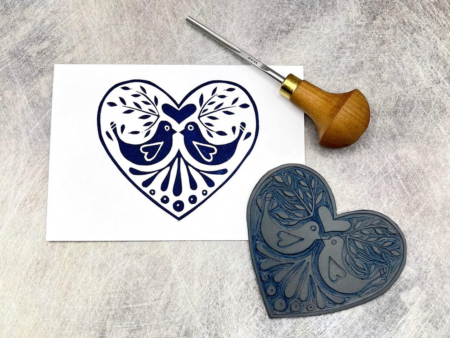 White card with an original lino printed blue heart design featuring two love birds, laid on a textured background. Photo includes lino and cutting tool used.