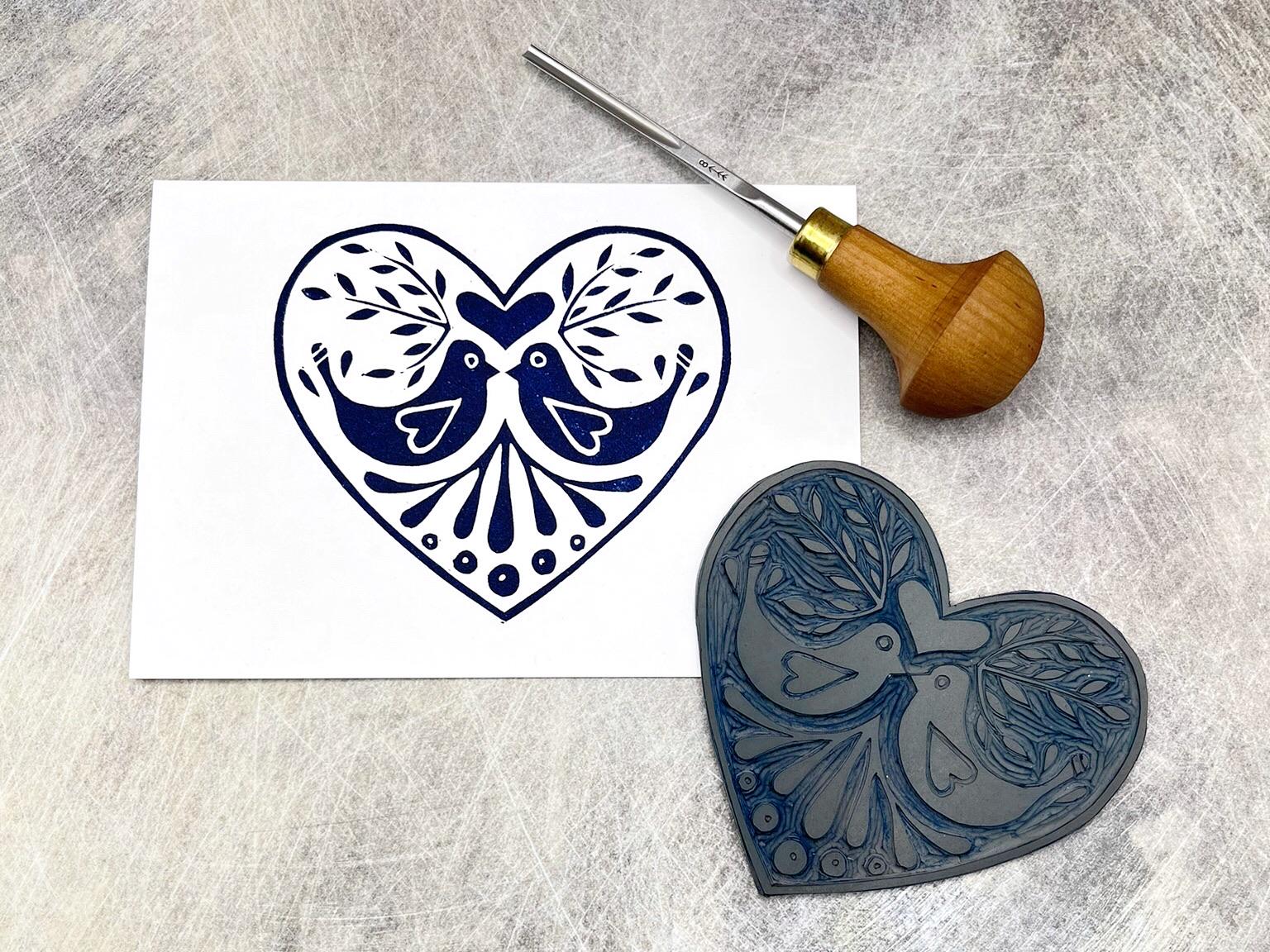 White card with an original lino printed blue heart design featuring two love birds, laid on a textured background. Photo includes lino and cutting tool used.