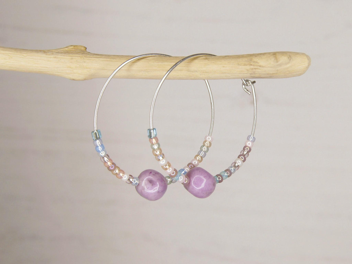 Close up of a pair of silver hoop earrings with lilac Jasper and seed beads hanging on driftwood against a light surface.
