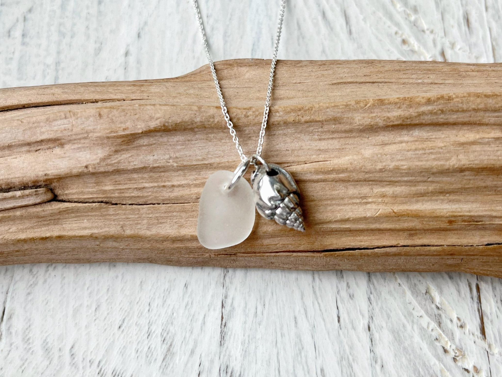 Necklace with white sea glass and silver shell pendant laid on driftwood against a textured surface
