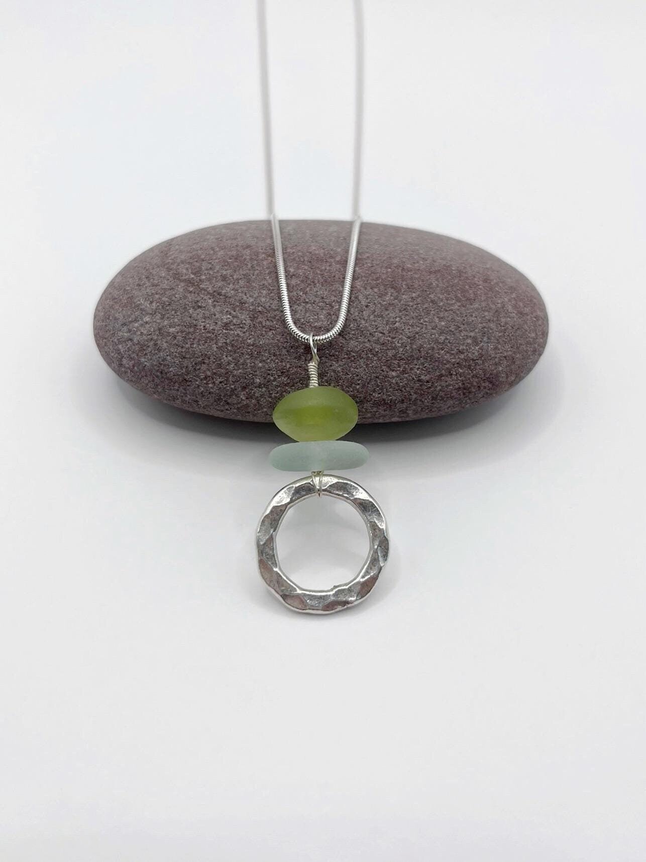Necklace with a silver ring pendant, jade bead and sea glass laid against a purple pebble on a white background