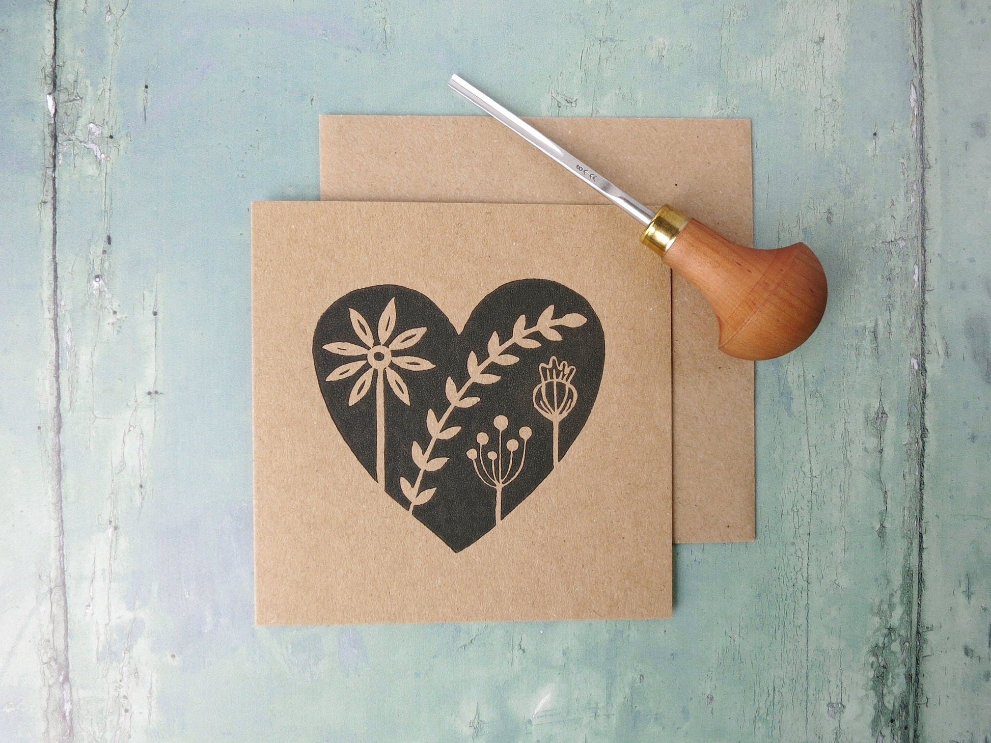 Kraft paper card with an original black lino printed heart design featuring floral elements laid on a textured surface. Photo includes lino cutting tool