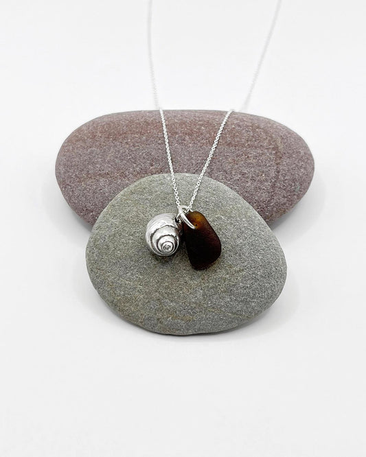 Silver necklace with burnt orange sea glass and a silver shell pendant laid on pebble against a white background