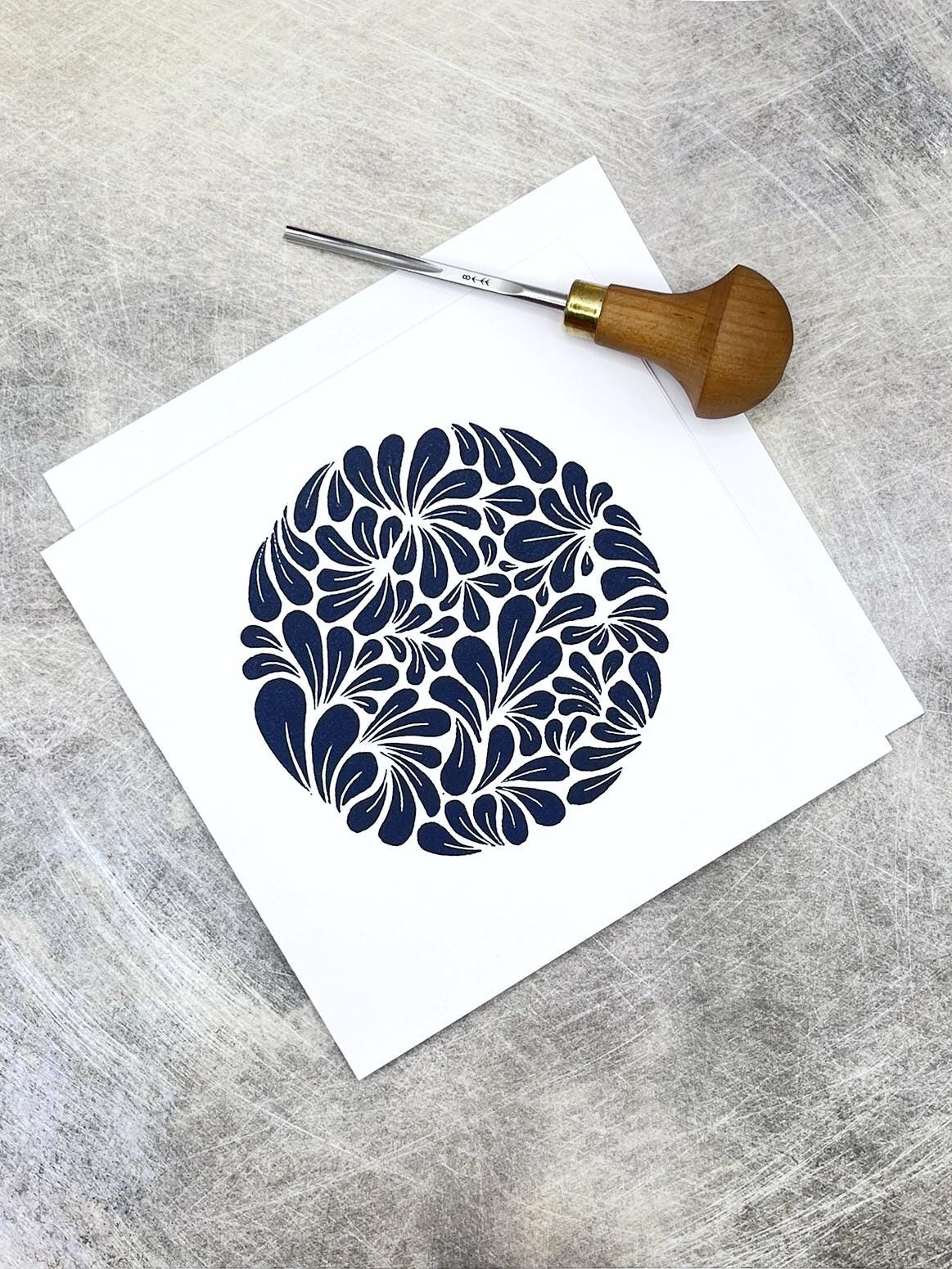 Original navy blue lino printed mussel shell pattern on white card laid on a textured background. Photo includes lino cutting tool