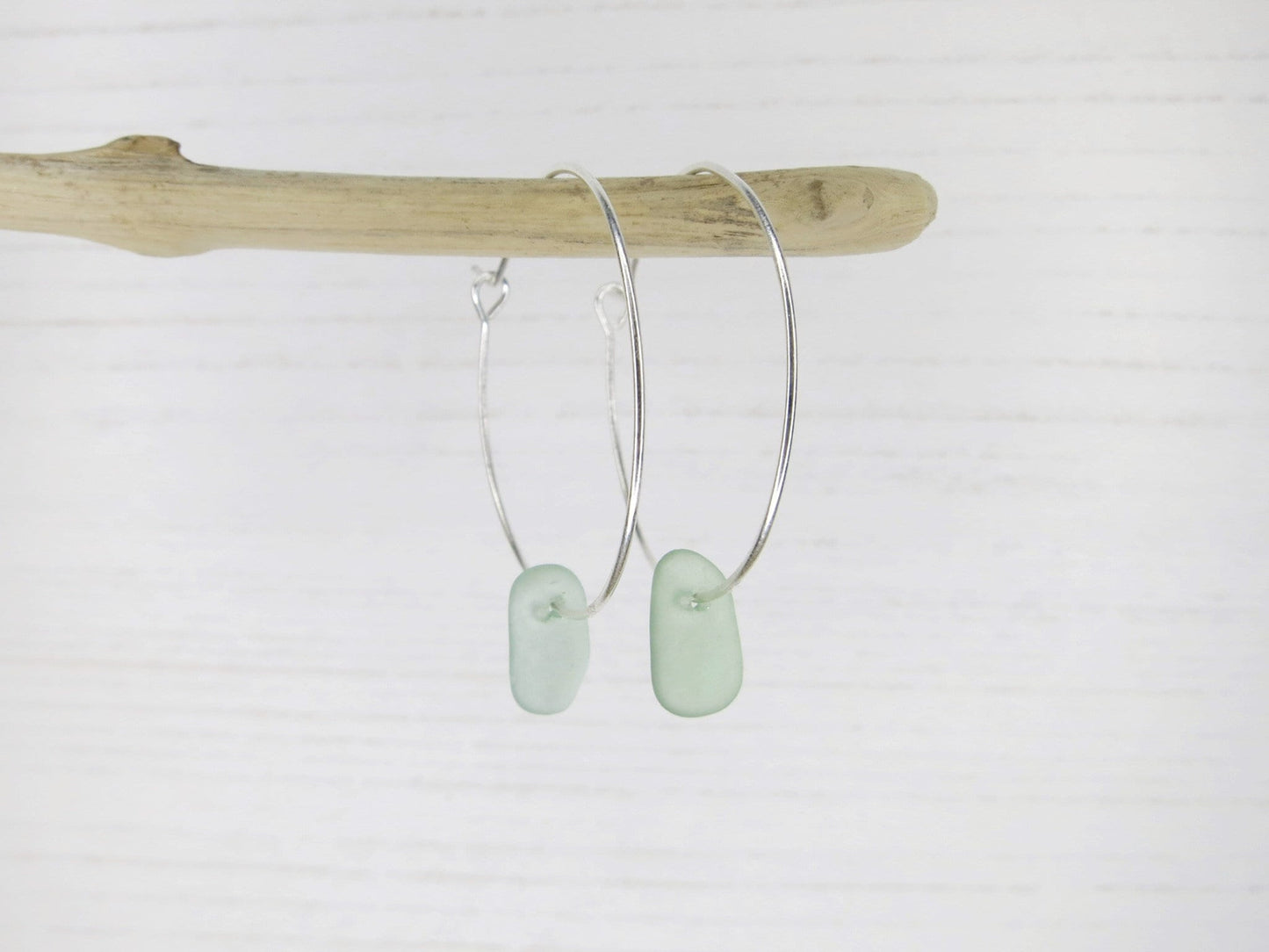 Pair of silver hoop earrings with sea foam coloured sea glass hanging on driftwood against a white background