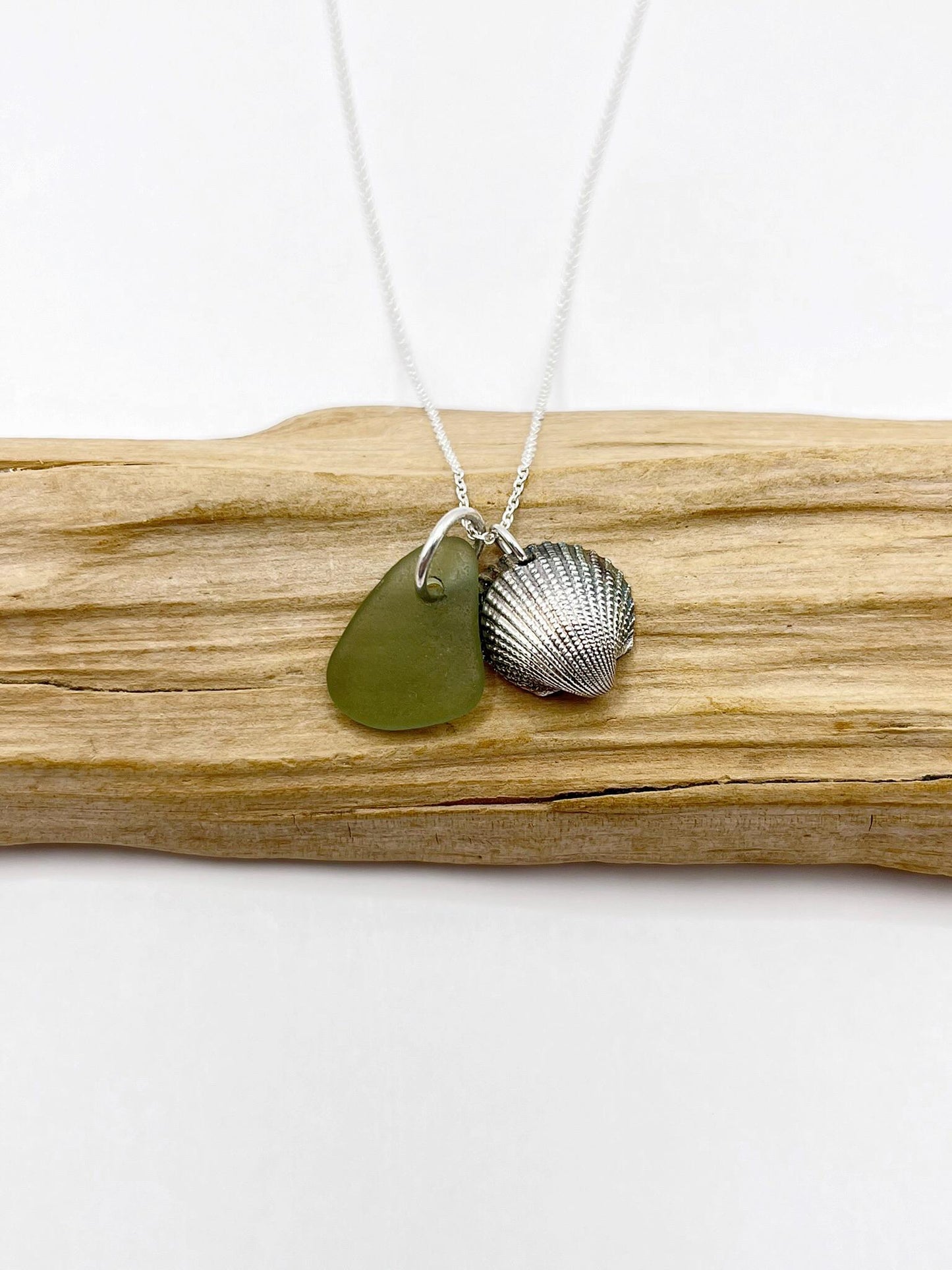 Necklace with green sea glass and a silver shell pendant laid on a piece of driftwood.