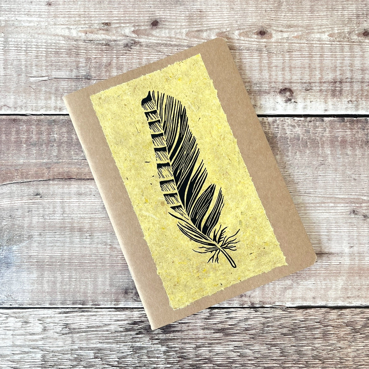 A5 kraft paper notebook or journal with a textured yellow handmade paper panel featuring a black lino ink feather illustration, photographed flat on a rustic wood plank surface.