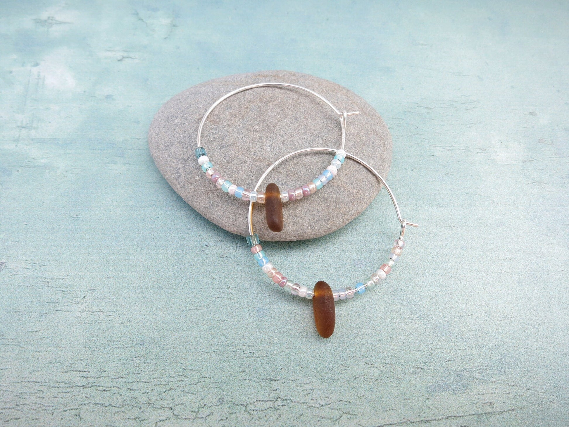Silver hoop earrings with pastel seed beads and burnt orange sea glass, laid on a grey pebble against a light blue background