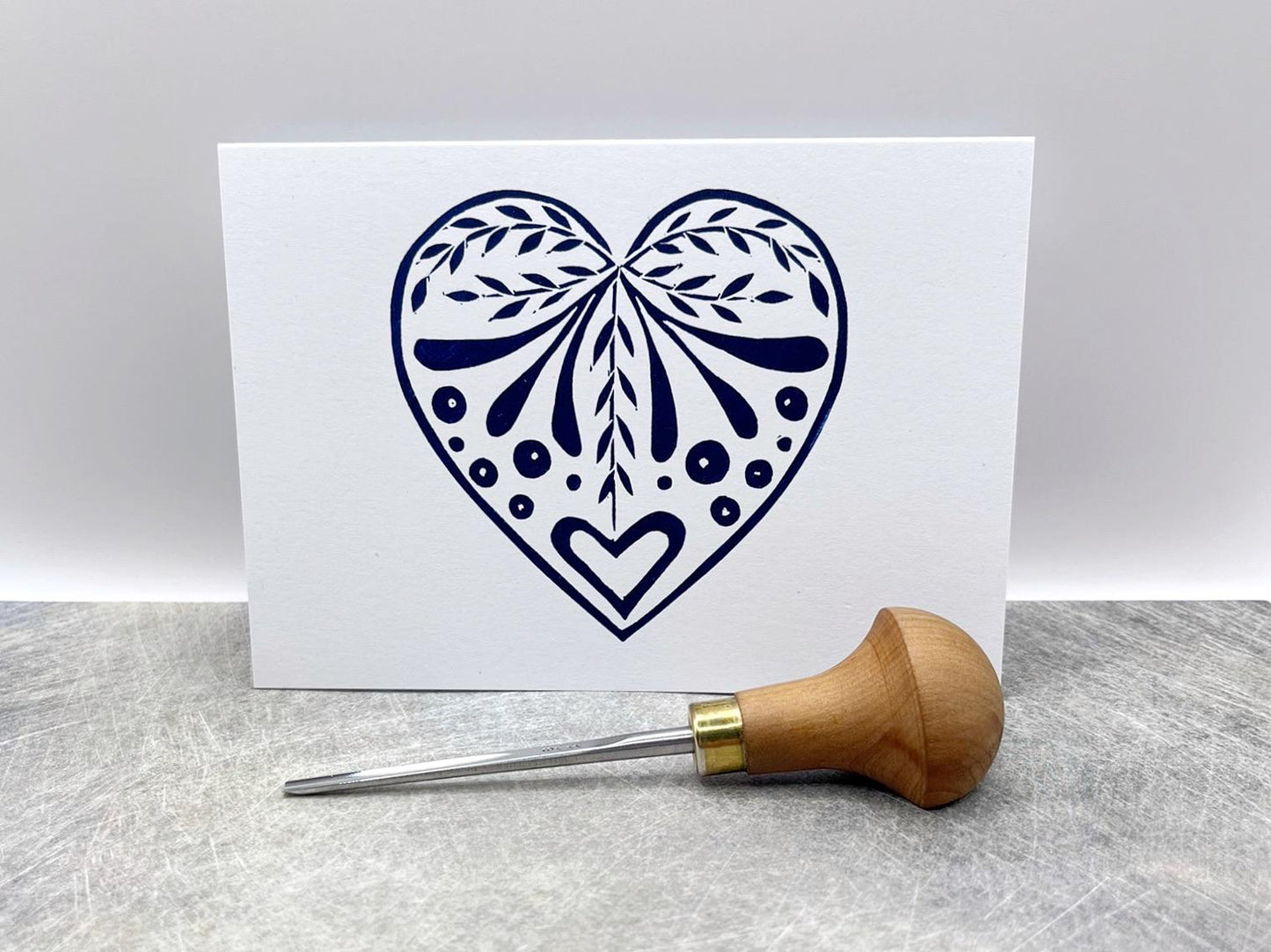 Blue heart card from the set of three white cards with original lino printed heart designs. Stood on a textured surface. Photo includes lino cutting tool.