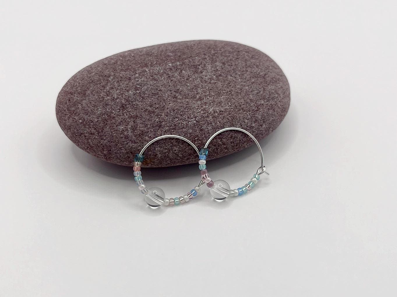 Pair of pastel seed bead and clear crystal hoop earrings laid against a purple pebble on a white surface