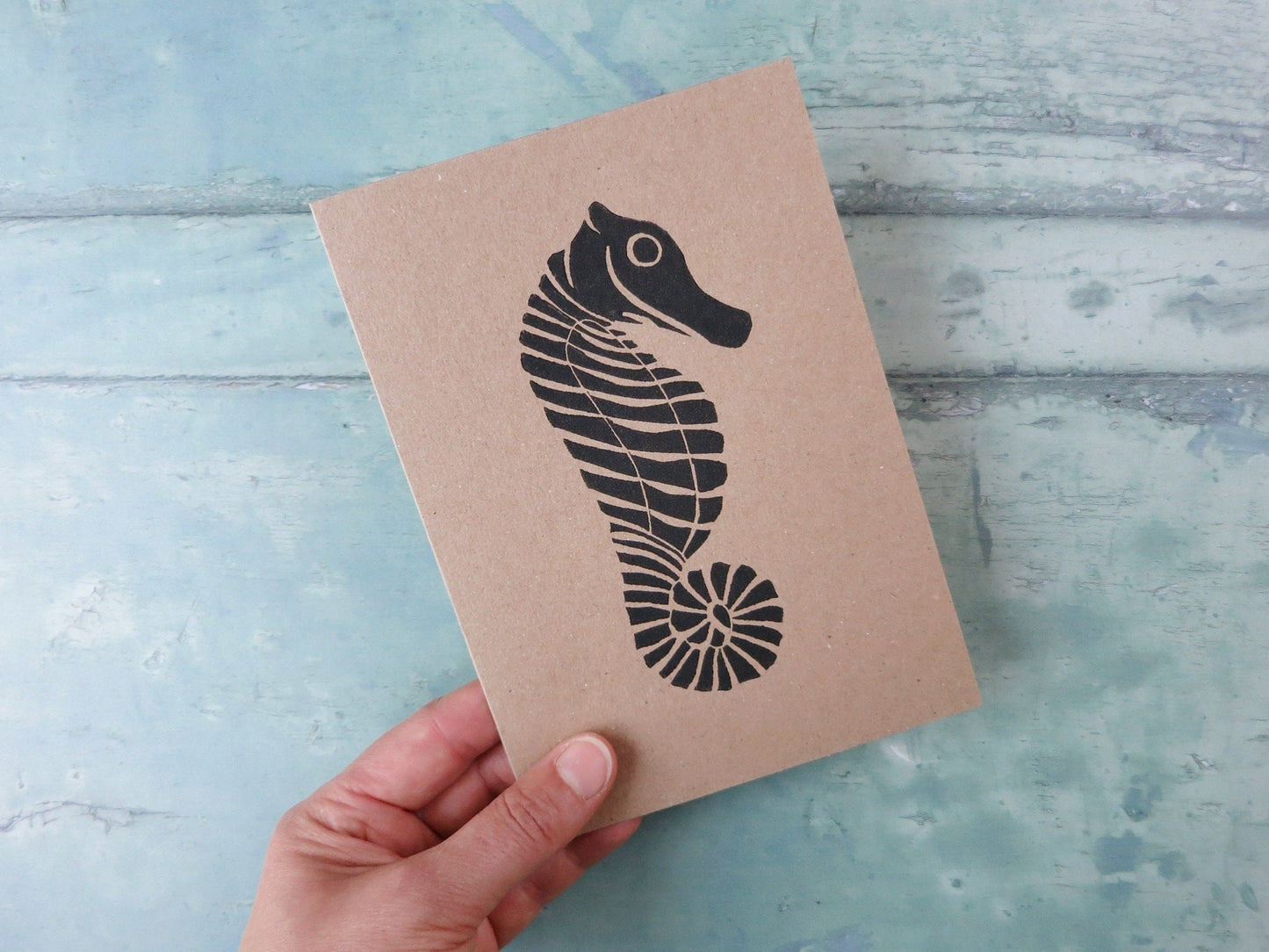 Hand holding Kraft paper card with an original seahorse lino print laid on a textured blue surface. 