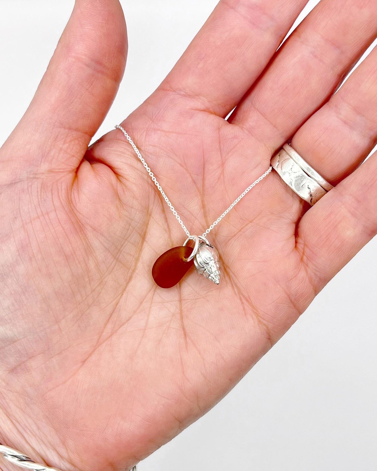 Necklace with burnt orange sea glass and silver shell pendant held in a hand against a white background