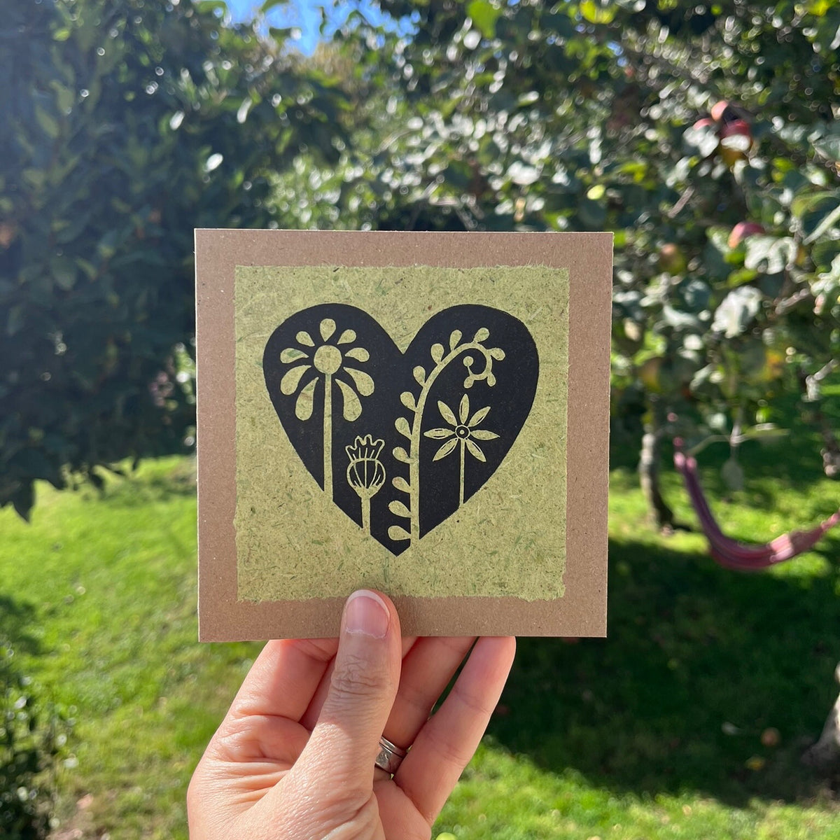 Hand holding a square kraft greeting card with a black floral heart lino print on pale green handmade paper mounted on the front. Held against a garden backdrop.