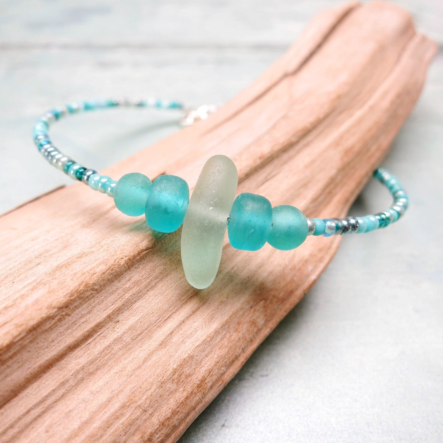 Close up of bracelet with pale sea foam sea glass and sea green and turquoise glass beads laid on driftwood against a pale blue background