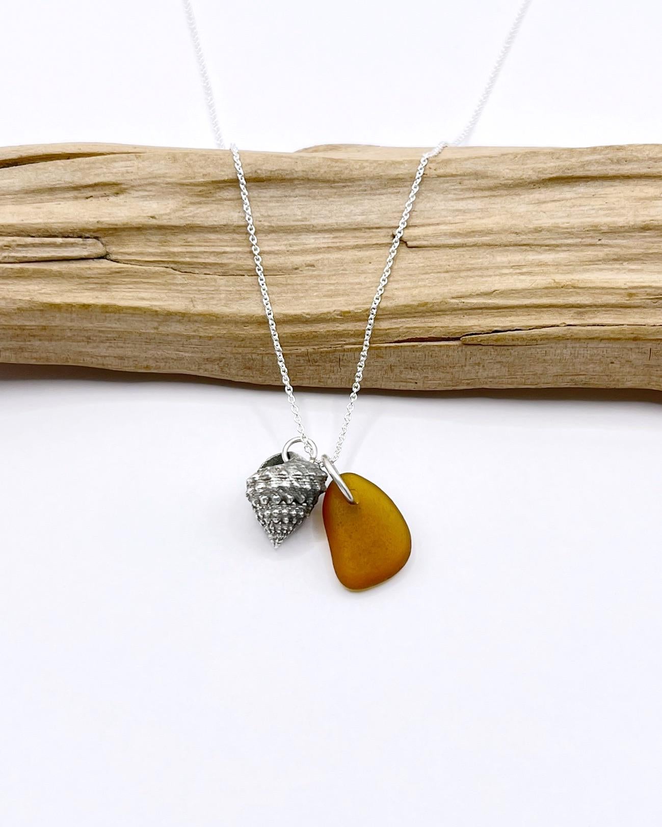 Necklace with burnt orange sea glass and silver shell pendant laid in front of driftwood against a white background