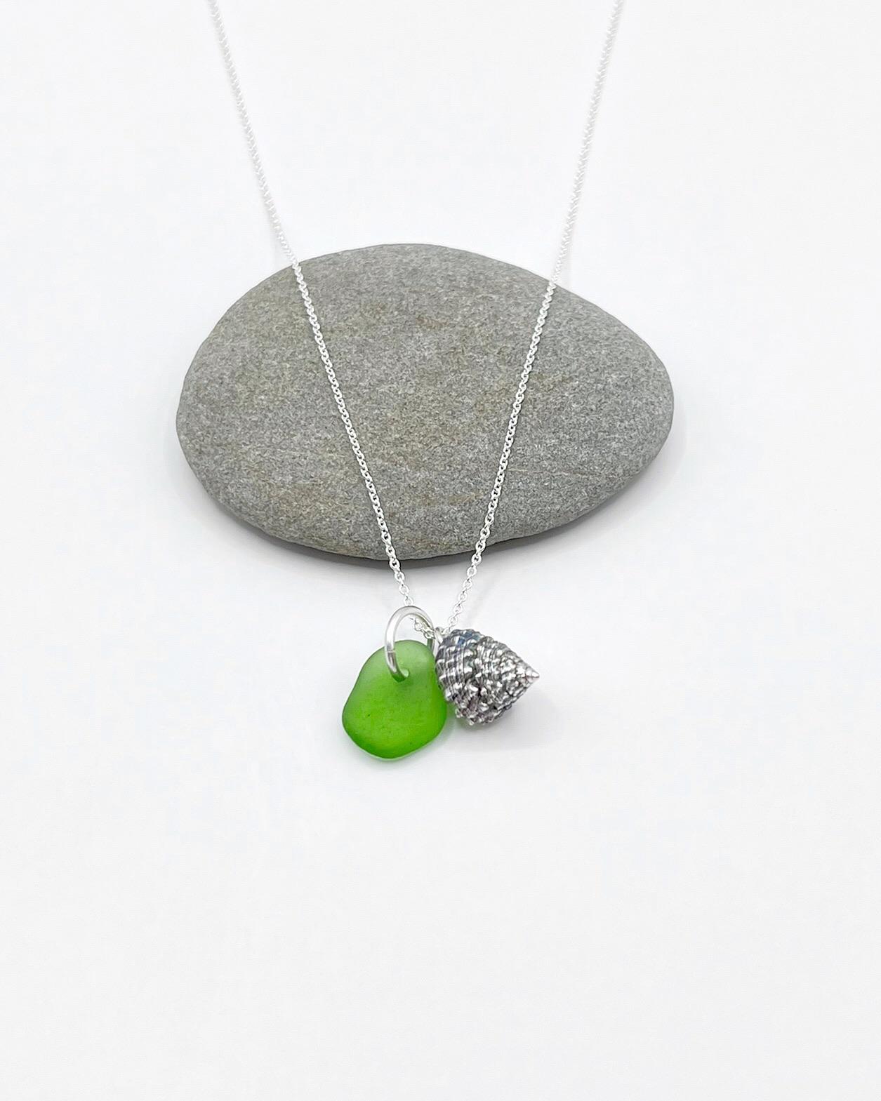 Necklace with green sea glass and silver shell pendant laid in front of a grey pebble on a white background