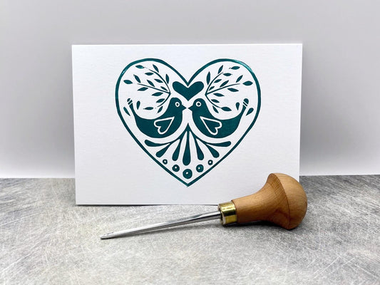 White card with an original lino printed green heart design featuring two birds. Photo includes lino cutting tool stood against a textured background.