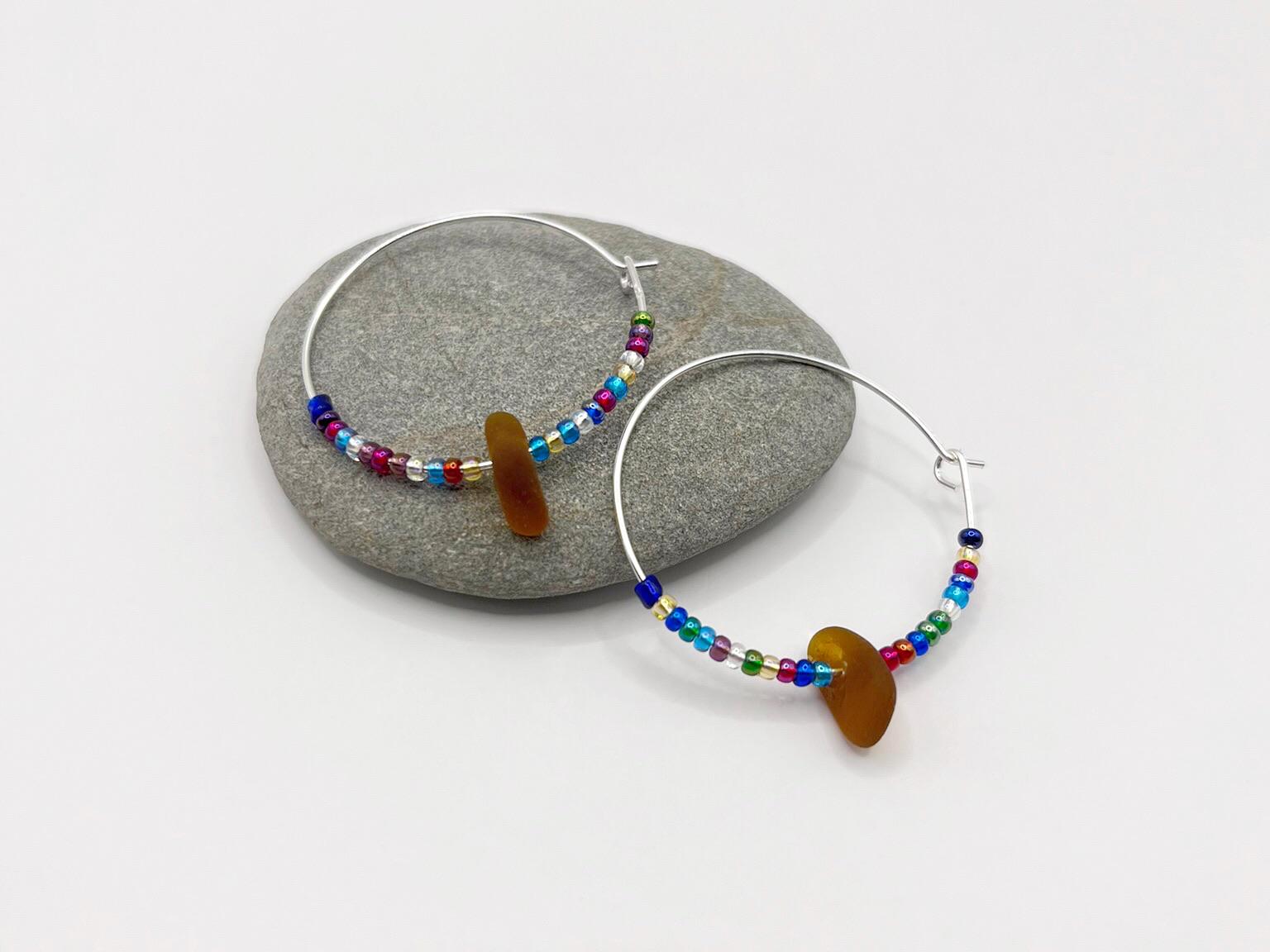 Rainbow seed bead and burnt orange sea glass hoop earrings laid on a grey pebble against a white background
