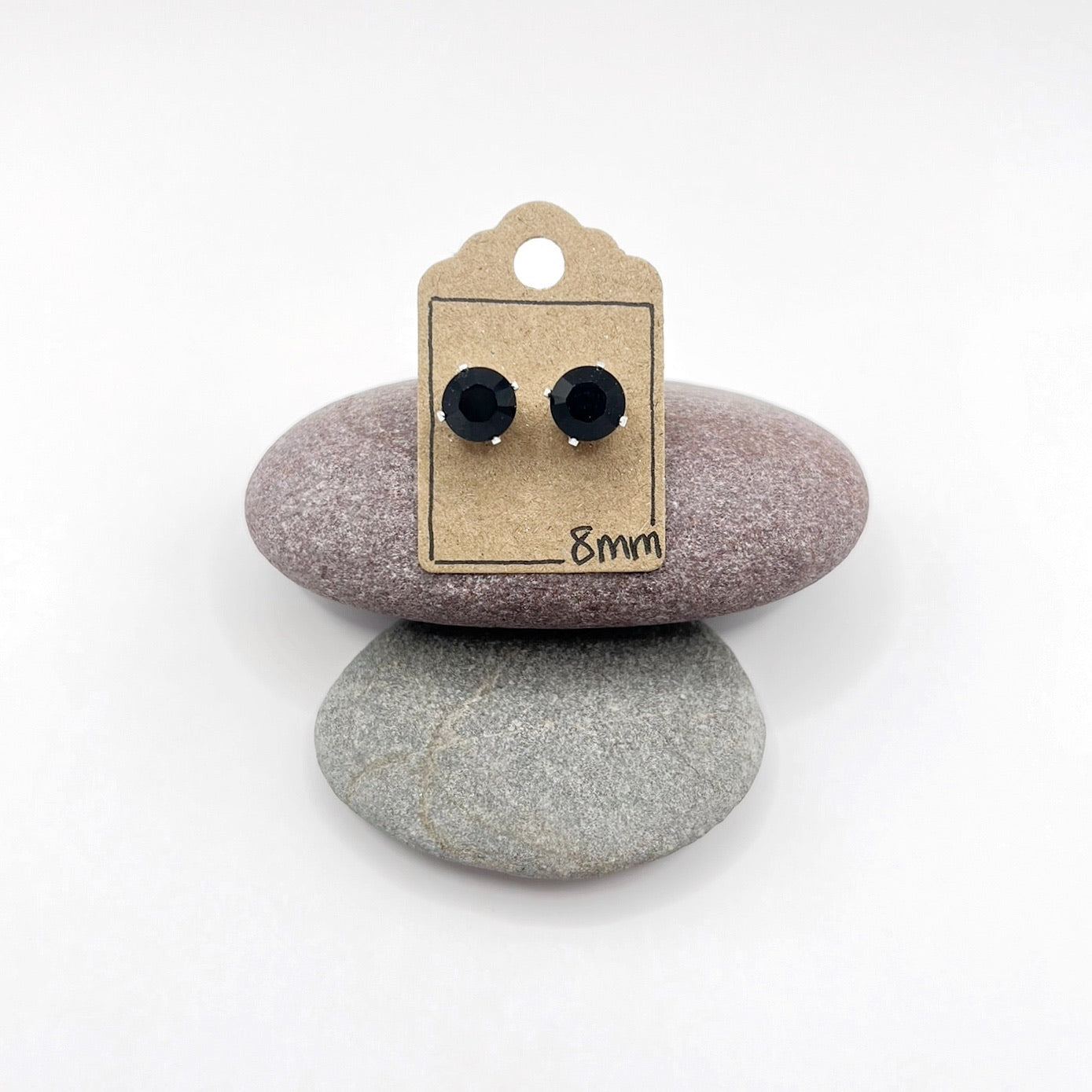 Black cut glass stud earrings on a cardboard backing with '8mm' text, placed on two stones.