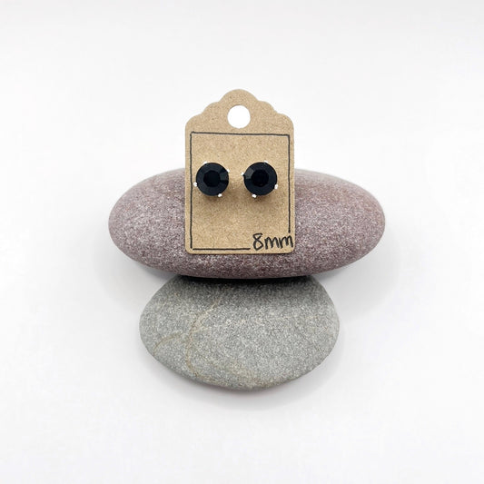 Black cut glass stud earrings on a cardboard backing with '8mm' text, placed on two stones.