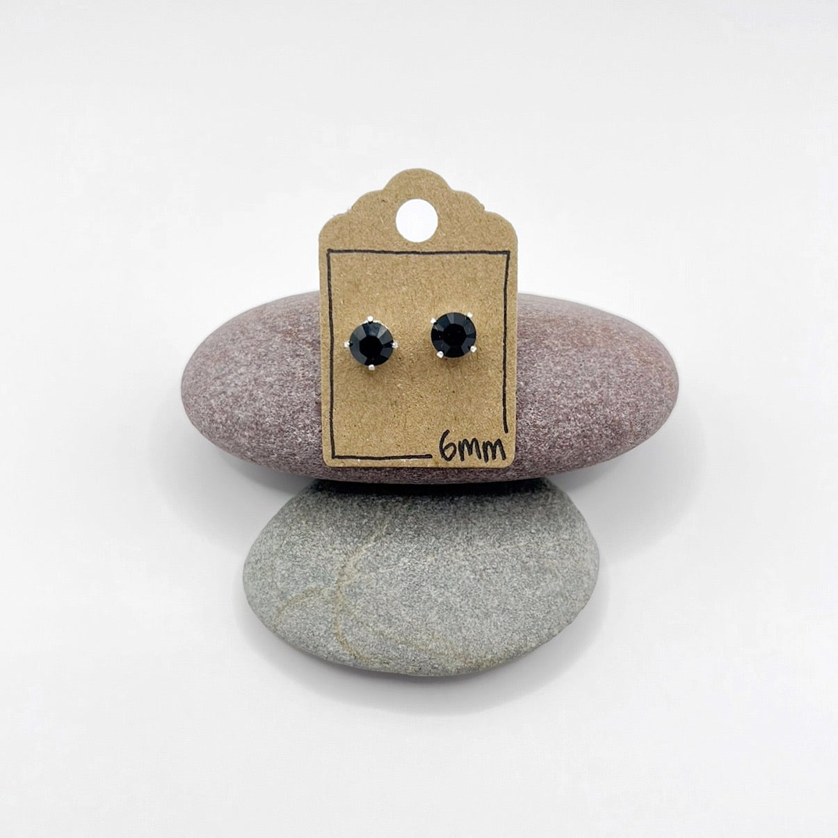Black stud earrings on a cardboard display card with '6mm' text, placed on two stones.