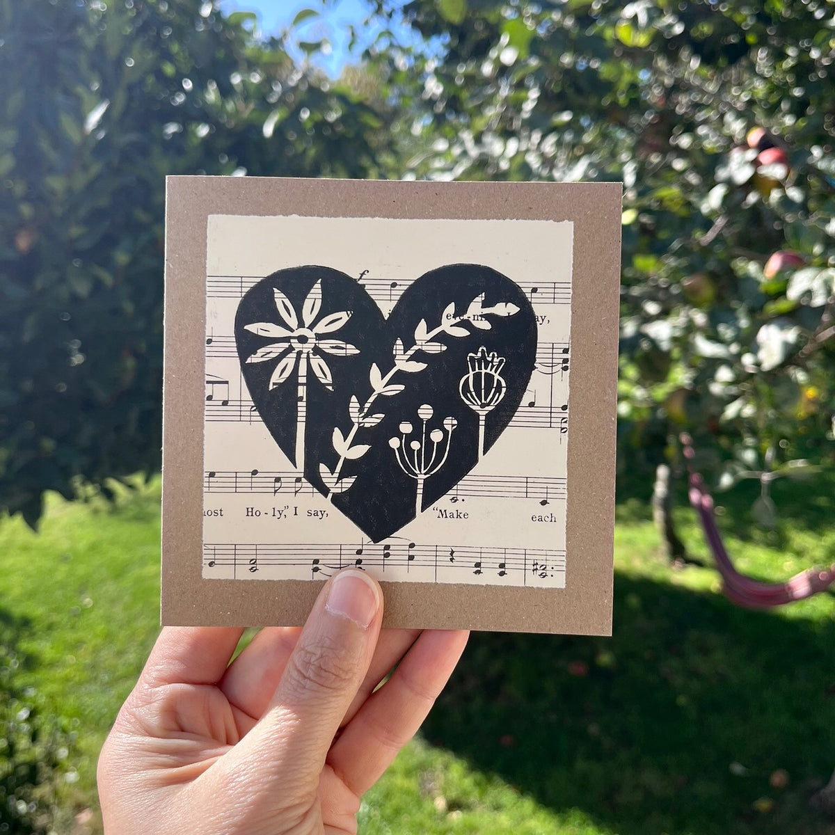 A hand holding a square kraft greeting card with a black floral heart lino print on vintage music paper mounted on the front. Held against a garden backdrop.