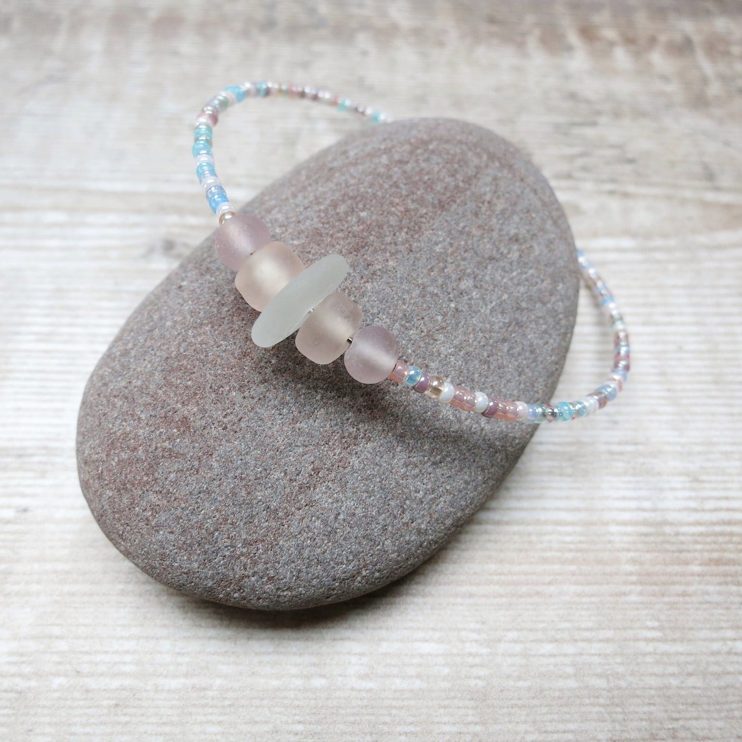 Bracelet with white sea glass and pastel beads laid on a purple pebble against a wooden background